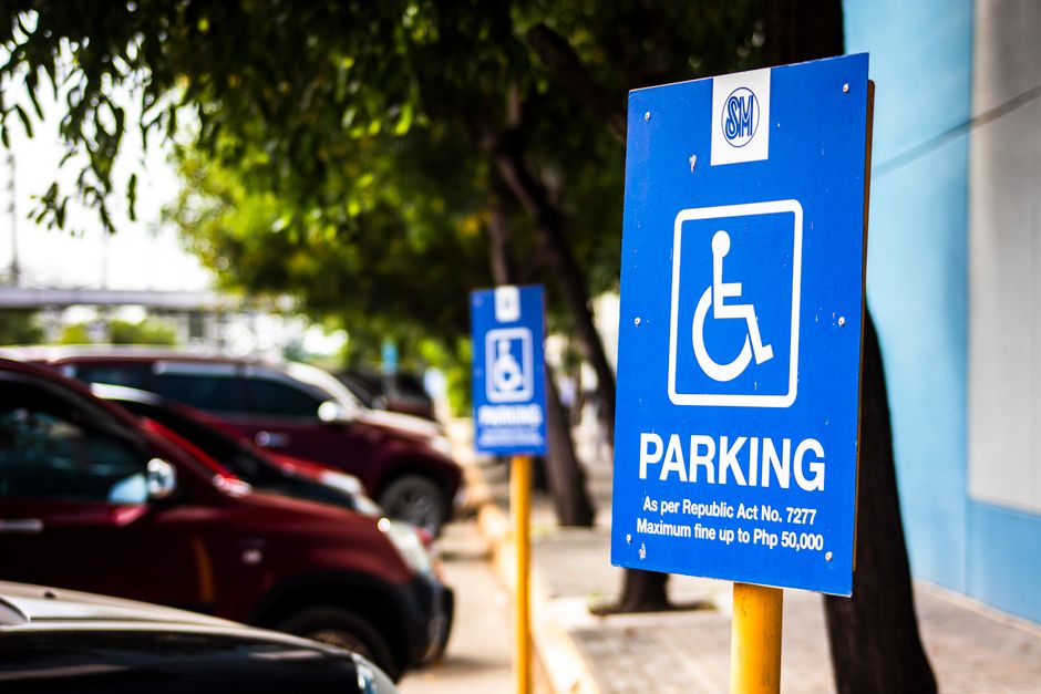 A circular no parking sign with a red border, blue background, and a red diagonal line across the center, mounted on a metal pole standing against a brick wall. The wall features a mix of beige and li