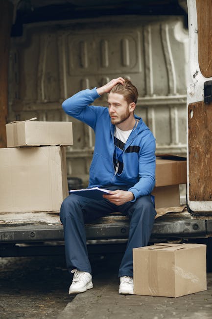A man with a beard, wearing a black cap and a white polo shirt, is standing outside near a moving van during daytime. He is holding a medium-sized cardboard box, which appears to be packed for a home