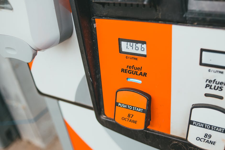 Close-up of a fuel pump at a petrol station showing a digital display reading 14.466 litres, with the fuel type labeled as 'refuel REGULAR'. The pump features an orange and white color scheme, with a black and orange 'PUSH TO START' button beneath the display, indicating octane levels (87 OCTANE). The surrounding area includes part of the nozzle handle on the left side, and the background suggests an outdoor setting with natural lighting. This image relates to home relocation services offered by Man with Van Hanworth, illustrating the importance of fuel management during furniture transport and packing and moving processes within house removals.