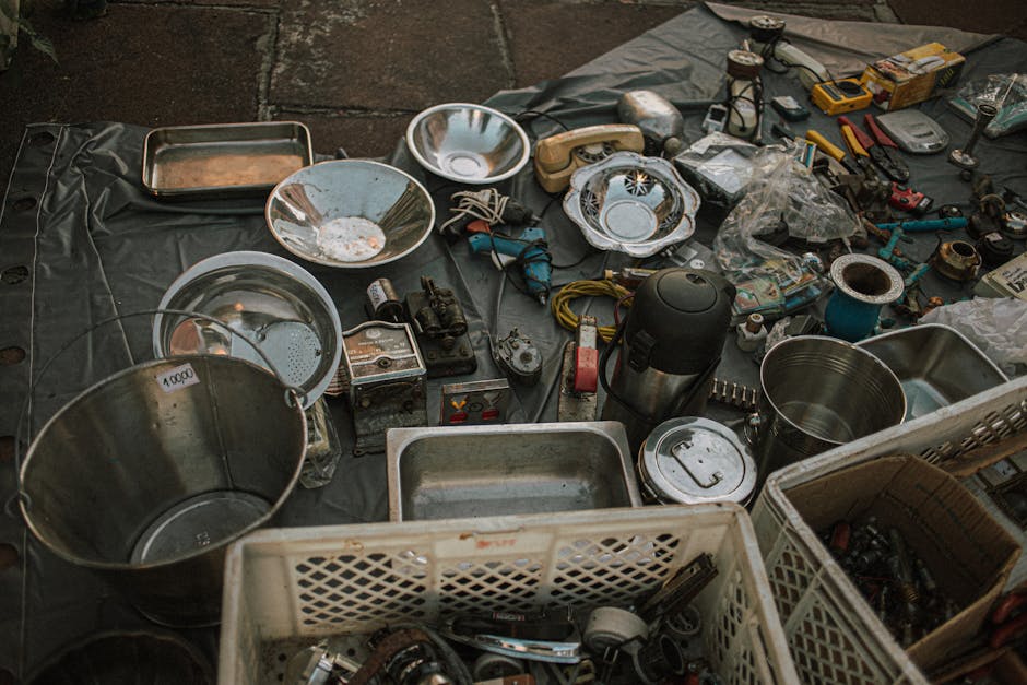 An arrangement of various household items and tools laid out on a black plastic sheet, including metal bowls, a stainless steel bucket, a plastic crate filled with miscellaneous objects, a thermos, a cardboard box with tools, wires, a cordless drill, bolts, and small hardware components. Some items are wrapped or partially covered, with others exposed, demonstrating a typical packing process during a house removal or home relocation. The scene appears to be set indoors or in a covered area, with any visible lighting providing a neutral tone. The assortment reflects preparations for furniture transport and efficient packing by a professional removals service such as Man with Van Hanworth, who handle bulky-item collections and moving logistics, including packing and loading activities.