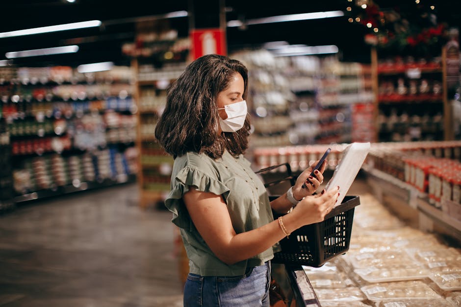 A woman wearing a medical face mask stands inside a hardware or home improvement store, holding a mobile phone in one hand and a white package or ledger in the other. She has shoulder-length brown curly hair and is dressed in a light green blouse with ruffled sleeves and blue jeans. Her shopping basket rests on a cart or trolley, positioned at her side. The store environment features wooden shelves stocked with various hardware supplies, containers, and packaged goods, with some items arranged on display racks behind her. The aisle floor is concrete, and bright store lighting illuminates the scene, emphasizing the rows of products and the woman's focused attention on her phone or the item she is holding. This setting illustrates the process of planning or organizing a home relocation, such as checking removal quotes or packing details, with Man with Van Hanworth providing furniture transport and moving logistics.