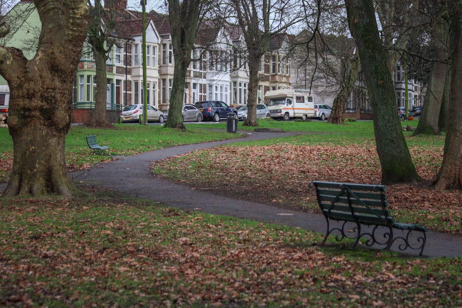 A residential street scene featuring a park area with several large mature trees and a winding paved pathway covered in fallen leaves, indicating an autumn setting. In the foreground, there are two black metal benches with wooden slats, positioned along the pathway, with one bench facing towards the viewer and the other further away. In the background, a row of Victorian-style terraced houses with bay windows and decorative facades are visible, along with parked cars along the street, including a small white van positioned near the curb. The street scene is well-lit with natural daylight, and the environment appears calm and organized. While no active moving or loading process is visible, this image reflects the typical environment where home relocation and furniture transport could take place, aligning with services offered by Man with Van Hanworth, especially in relation to parking, access, and timing considerations for house removals.