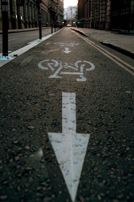 A close-up view of a dedicated bike lane on a city street during daytime, with white painted symbols and directional arrows on the asphalt indicating bicycle traffic flow. The lane is separated from vehicle lanes by small black posts with reflective strips, and is bordered by tall buildings with large windows and stone facades on both sides. In the background, traffic lights and streetlights are visible, along with some parked vehicles, suggesting an urban environment suitable for home relocation or moving services. The street surface appears textured, and the scene is lit with natural daylight, creating a clear and detailed environment consistent with urban moving logistics and transport processes.