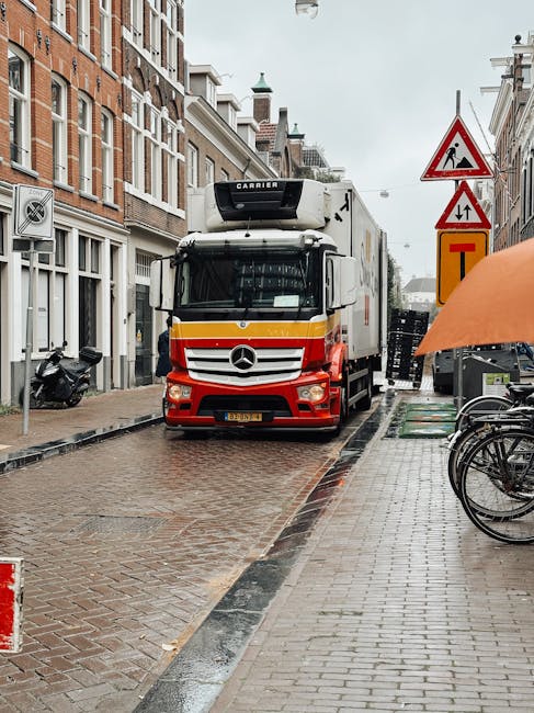 A large delivery truck with a white cab and a Mercedes-Benz logo on the front is parked on a narrow cobbled street in Hanworth, adjacent to residential buildings. The truck appears to be involved in a home relocation, with its rear doors possibly open for loading or unloading furniture and boxes. The street shows typical urban features, including traffic signs warning of roadworks and lane restrictions, with a partially visible orange umbrella and bicycles parked on the pavement. The surrounding buildings are multi-storey, constructed from brick, with some architectural detailing, and the sky above is overcast, indicating rainy weather. The scene captures the logistical process of furniture transport and packing during a house removal, where careful manoeuvring of bulky items is necessary within confined street spaces, and [COMPANY_NAME] is likely assisting with the loading process.