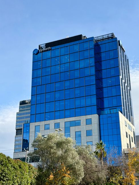 Photograph of a modern high-rise office building with a predominantly blue glass façade, reflecting the sky and surrounding structures. The building has multiple levels with a combination of glass pan