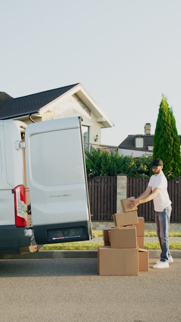 A young man dressed in casual clothing is engaged in a home relocation process, standing on a paved street next to a white moving van with its rear doors open. Inside the van, visible protective padding and cardboard boxes are arranged, ready for loading. Outside, he is stacking unpacked cardboard boxes onto a small pile on the pavement, handling one box with both hands. The boxes vary in size and are made of plain brown cardboard, some sealed with packing tape. The van is parked in front of a residential property with a well-maintained garden and a wooden fence, suggesting a suburban neighbourhood. Bright daylight and clear weather provide good natural lighting, highlighting the equipment such as trolleys and blankets used for safe furniture transport. The scene captures the logistics of packing and loading that are typical in professional removals, emphasizing the careful handling of household items during a relocation, as practiced by companies like Man with Van Hanworth, in response to city permits for moving vans in Hanworth.