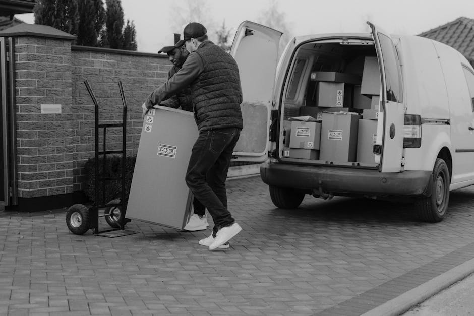 Two movers from Man with Van Hanworth are engaged in a home relocation task outside a property, loading a large cardboard box onto a hand truck. The movers are dressed in casual clothing with one wearing a cap and both wearing jackets, indicating outdoor working conditions. They are transferring items from the front door area onto a commercial white van parked on a paved driveway. The van’s rear doors are open, revealing numerous boxes and packages ready for transport, some secured with plastic wrapping or packing materials. The loading process involves careful handling of furniture and boxes into the van, with the movers coordinating to ensure safe and efficient packing. The background includes a brick wall and residential features, consistent with a typical house move in Hanworth, supported by a local removals service such as Man with Van Hanworth, providing house removals and furniture transport logistics.