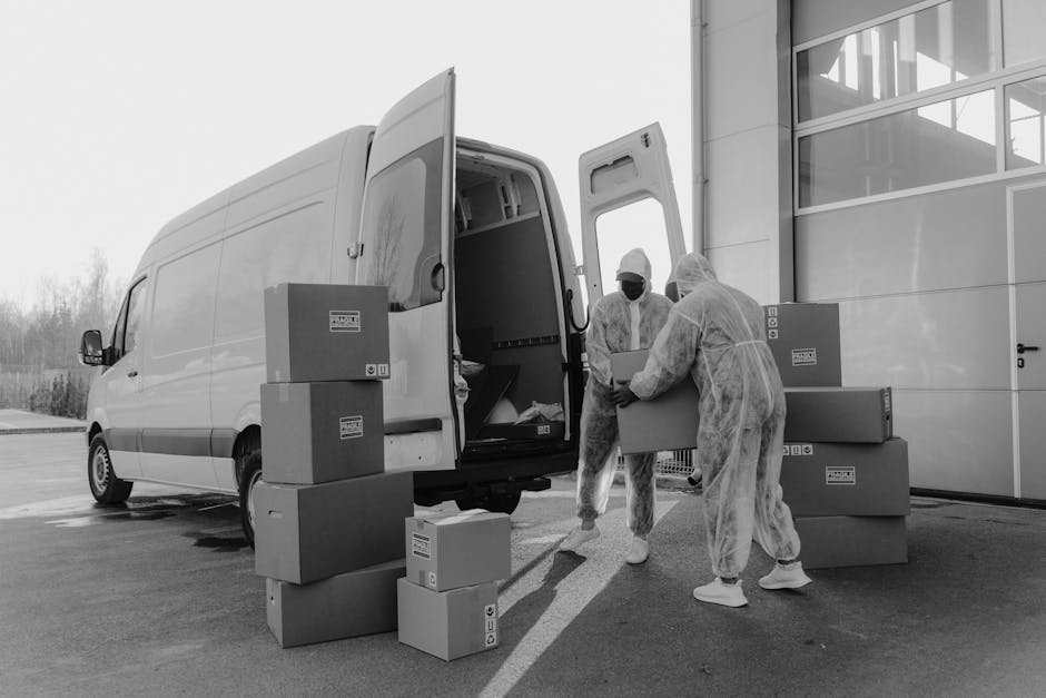 A young man dressed in casual clothing is engaged in a home relocation process, standing on a paved street next to a white moving van with its rear doors open. Inside the van, visible protective padding and cardboard boxes are arranged, ready for loading. Outside, he is stacking unpacked cardboard boxes onto a small pile on the pavement, handling one box with both hands. The boxes vary in size and are made of plain brown cardboard, some sealed with packing tape. The van is parked in front of a residential property with a well-maintained garden and a wooden fence, suggesting a suburban neighbourhood. Bright daylight and clear weather provide good natural lighting, highlighting the equipment such as trolleys and blankets used for safe furniture transport. The scene captures the logistics of packing and loading that are typical in professional removals, emphasizing the careful handling of household items during a relocation, as practiced by companies like Man with Van Hanworth, in response to city permits for moving vans in Hanworth.