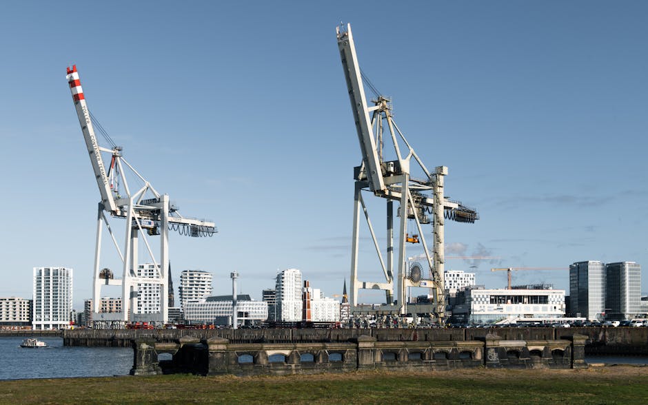 A wide view of a port area featuring two large industrial cranes with white and red striped arms and tall, metallic structures used for lifting and moving heavy cargo. The cranes are positioned on a concrete quay near the water, with a small boat sailing on the river in the foreground. In the background, there are modern white and grey buildings and high-rise apartments, with a clear blue sky overhead. This scene illustrates key elements of intermodal freight handling, which could relate to the logistics involved in house removals and furniture transport during a home relocation, as handled by companies like Man with Van Hanworth, situated in an urban setting with construction cranes visible in the distance.