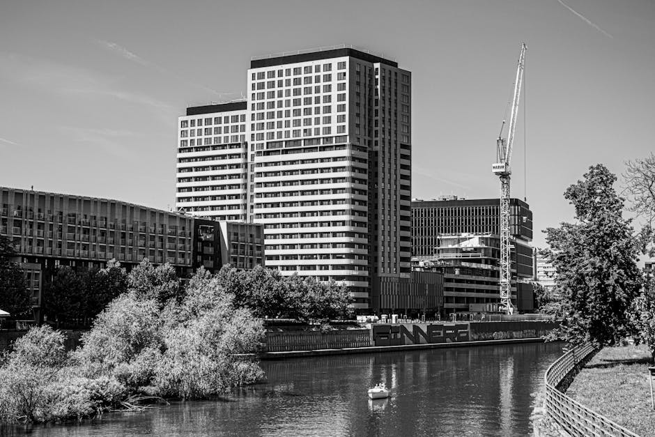 Black and white photograph of a modern urban scene featuring a large high-rise building with multiple floors and a grid-like window pattern, situated next to a river or canal. In the foreground, there are several lush trees with dense foliage along the water's edge, partially obscuring some lower structures. A construction crane is visible to the right of the high-rise, and nearby, there are contemporary office or apartment buildings with flat roofs and glass facades, suggesting ongoing development or renovation. The scene appears to be daylight with clear skies, and the water reflects the surrounding buildings and trees. The image is related to home relocation and furniture transport, illustrating an urban environment where professional removals, such as those provided by Man with Van Hanworth, might assist with moving logistics, packing, and loading procedures in a city setting.