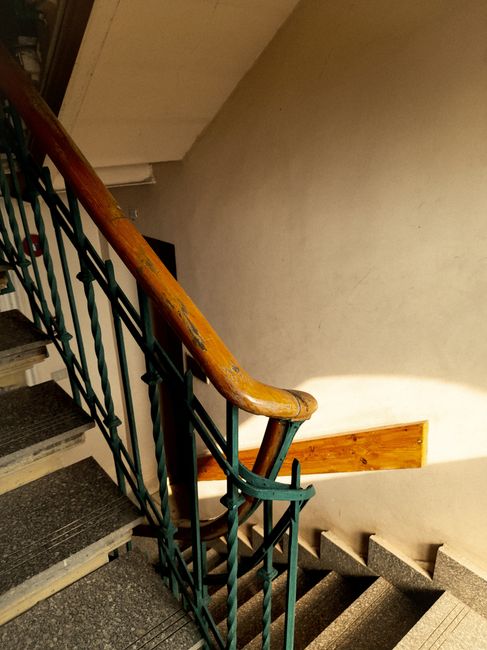 The image depicts an indoor staircase within a residential building, showing a wooden handrail and a metal railing painted in dark green. The staircase has many steps made of grey stone or concrete, with some steps being partially visible as they descend out of view. The wall alongside the staircase is painted in a light cream colour, with a rectangular wooden panel mounted on it. The lighting appears natural or coming from an overhead source, casting a soft glow on the wall. This setting is representative of typical stairwells encountered in flats or apartments, where space constraints often require careful navigation during home relocations. The image suggests a scenario involving furniture transport or moving logistics within a building, with the focus on the narrow, enclosed stairs that may necessitate specialized handling. The presence of the stairwell aligns with the topic of safe removal tactics for tight spaces, as performed by professional removal services such as Man with Van Hanworth.