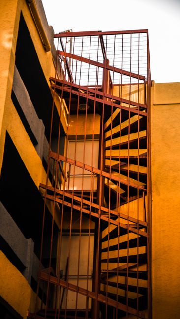 A multi-flight metal staircase painted in orange, located inside a building with yellow walls, leading upwards through a narrow, enclosed space. The staircase has a vertical and horizontal railing design, with the steps made of perforated metal. The lighting appears to be natural, coming from the top of the staircase, illuminating the metal framework and surrounding walls. In the context of house removals, this image illustrates a challenging feature of moving household items through confined stairwells typical of Hanworth flats. Professionals such as Man with Van Hanworth, specialising in removals, would need to employ careful packing, disassembling bulky furniture, and using appropriate lifting techniques to navigate this staircase during a home relocation or furniture transport process, especially in tight stairwell environments requiring safe and efficient removal tactics.