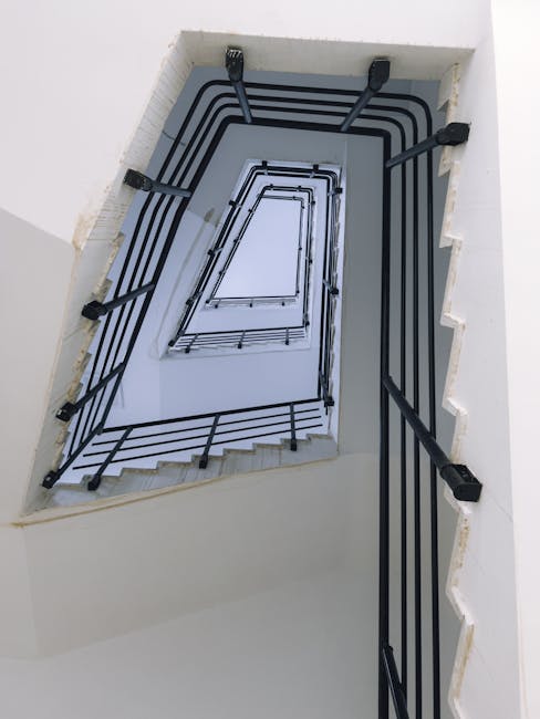 The image depicts an indoor staircase within a residential building, showing a wooden handrail and a metal railing painted in dark green. The staircase has many steps made of grey stone or concrete, with some steps being partially visible as they descend out of view. The wall alongside the staircase is painted in a light cream colour, with a rectangular wooden panel mounted on it. The lighting appears natural or coming from an overhead source, casting a soft glow on the wall. This setting is representative of typical stairwells encountered in flats or apartments, where space constraints often require careful navigation during home relocations. The image suggests a scenario involving furniture transport or moving logistics within a building, with the focus on the narrow, enclosed stairs that may necessitate specialized handling. The presence of the stairwell aligns with the topic of safe removal tactics for tight spaces, as performed by professional removal services such as Man with Van Hanworth.