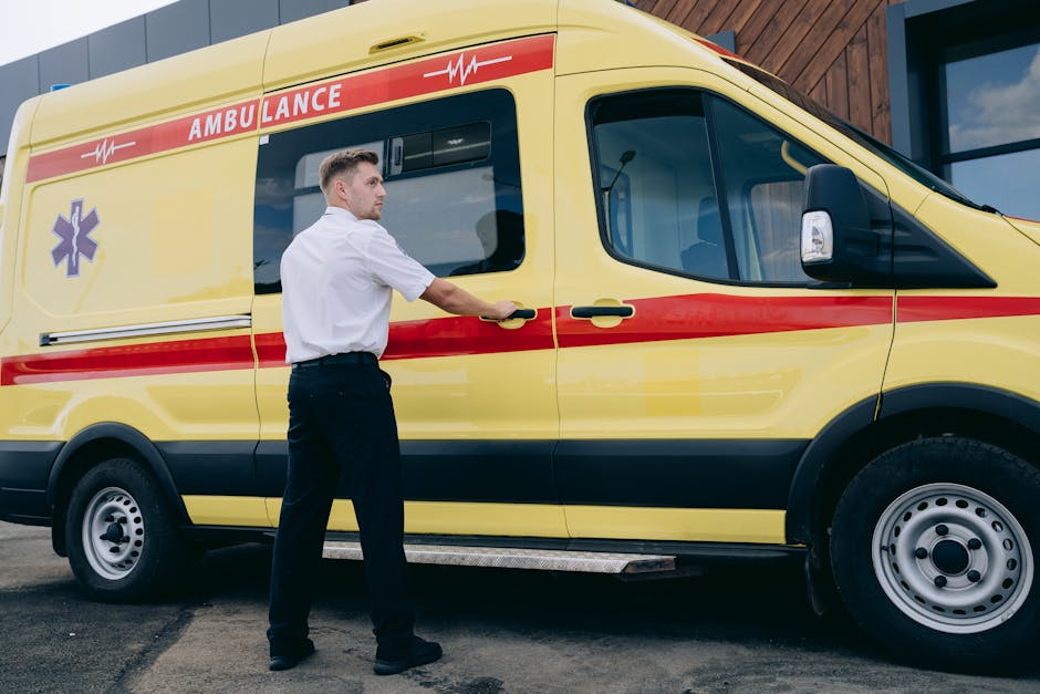 A male emergency medical technician wearing a white uniform shirt and black trousers is standing outside next to a yellow ambulance with red and purple markings. The technician is holding the ambulance's side door handle, preparing to open it. The ambulance is parked on a paved area adjacent to a modern building with large glass windows and wooden paneling. The vehicle's side windows reflect the surrounding environment, and the overall scene suggests readiness for a home relocation or transport service involving specialist transportation of individuals. Man with Van Hanworth occasionally delivers similar moving logistics and vehicle loading services as part of their removals operations.